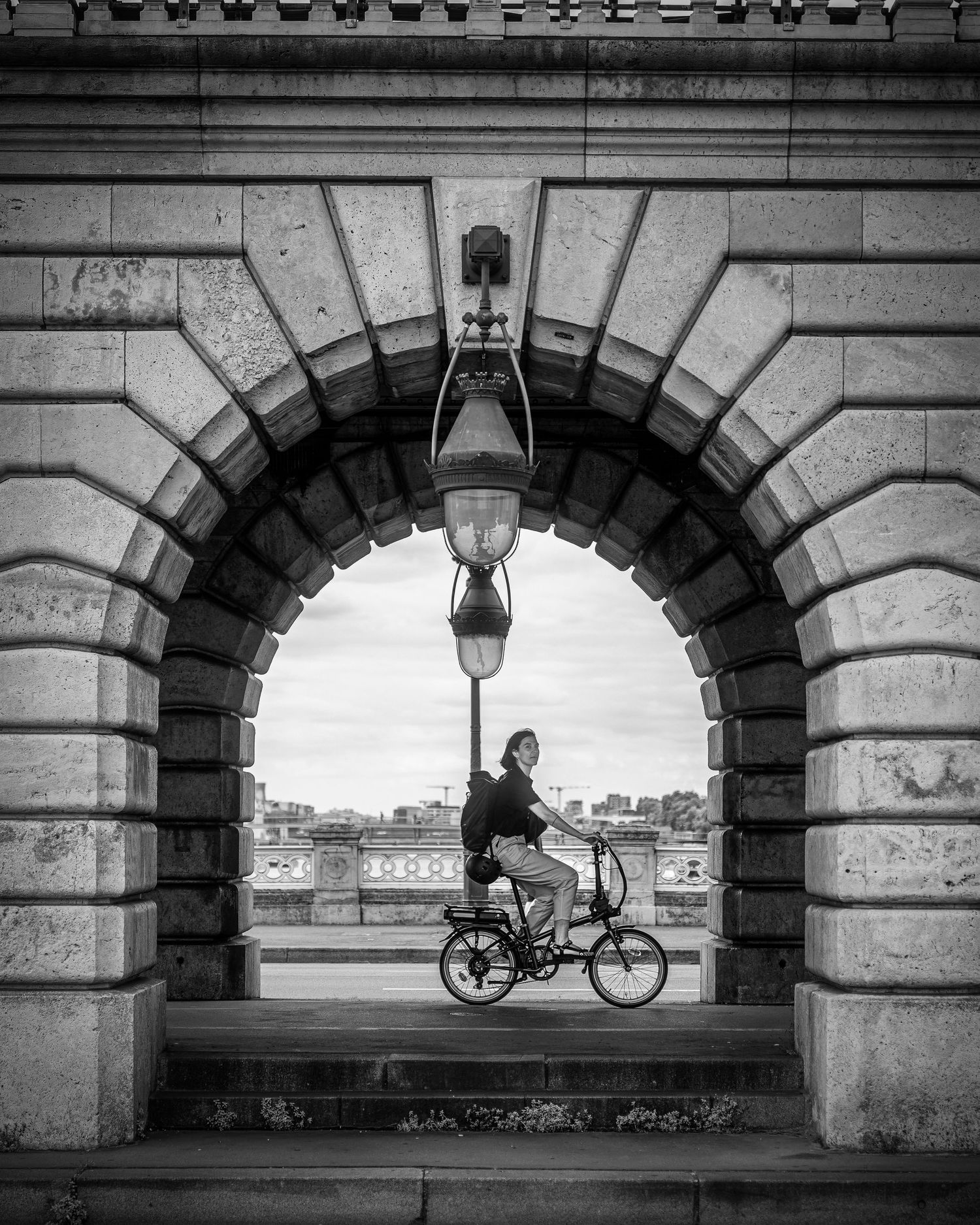 Black and white photo of a cyclist riding through a stone arch bridge with a vintage lantern overhead.