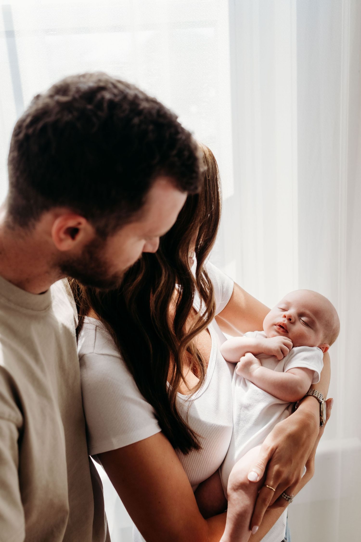 Young parents lovingly gazing at their newborn baby, mother holding infant in white onesie near bright window.