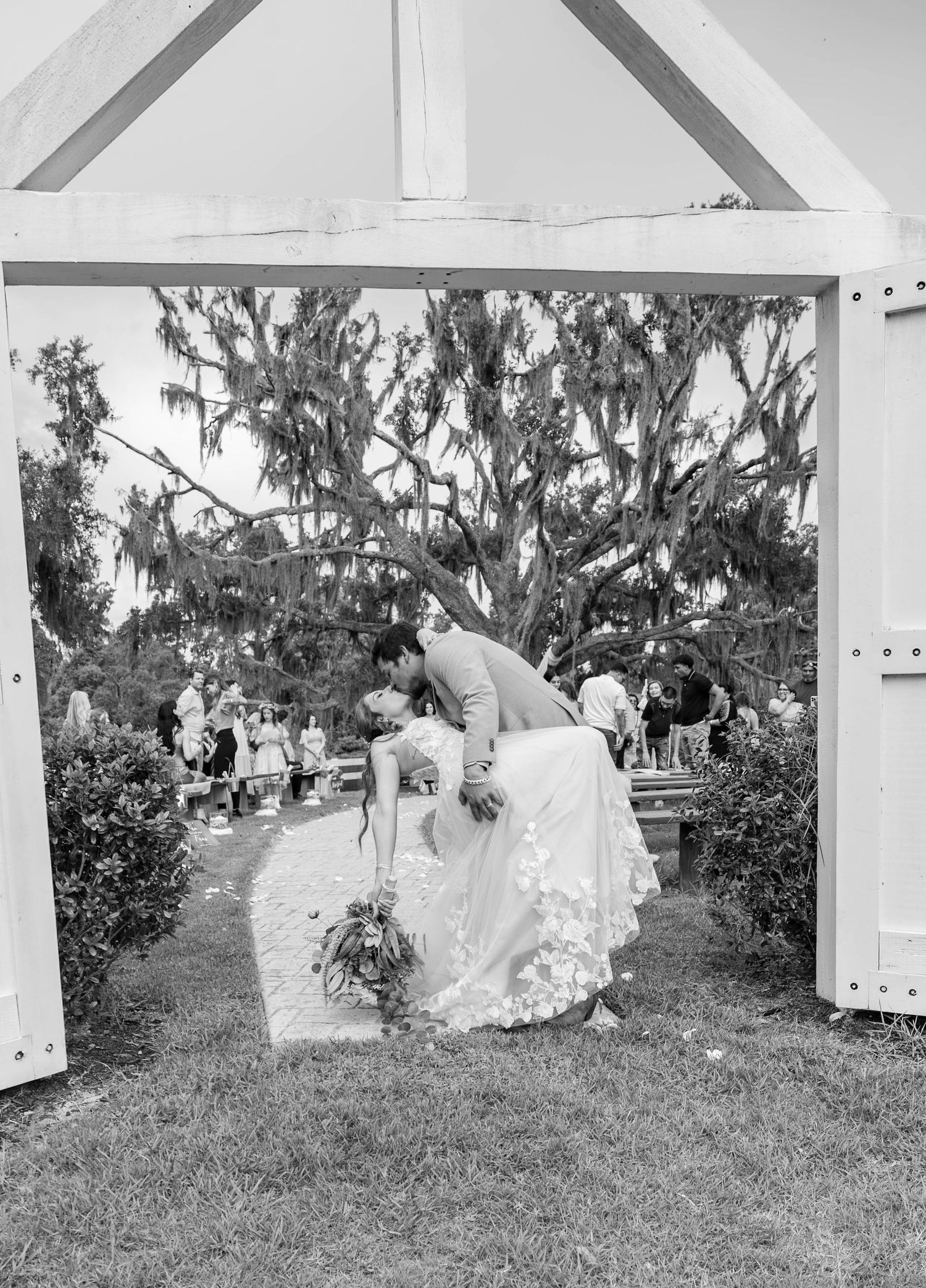 Black and white photo of bride and groom sharing a kiss under a white arch at outdoor ceremony.