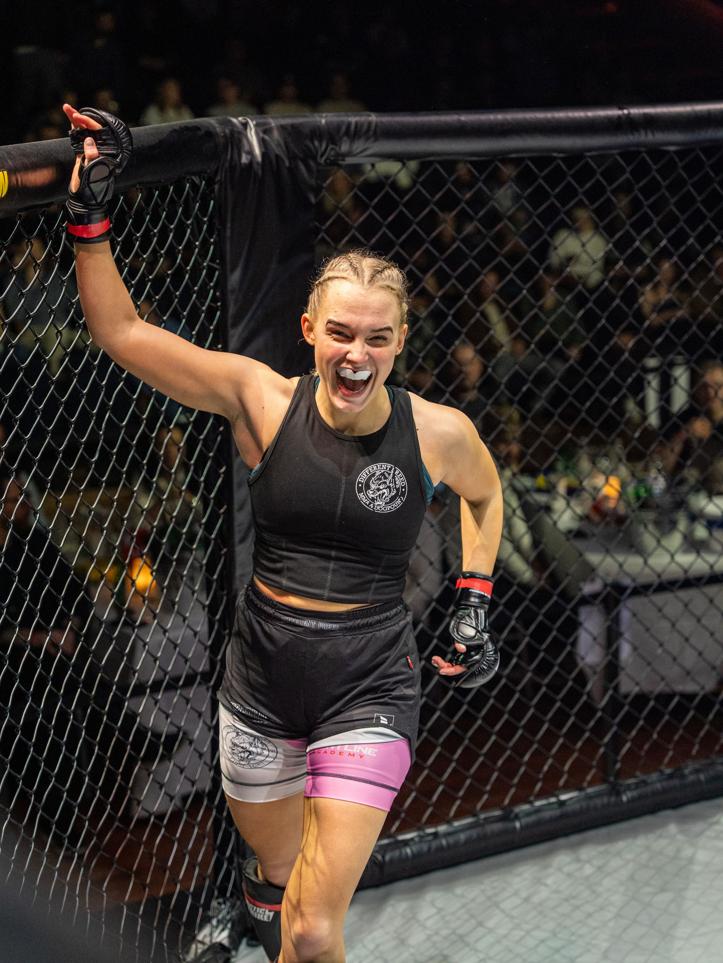 Female MMA fighter celebrates victory in cage, raising fist triumphantly, wearing black tank top and pink shorts.