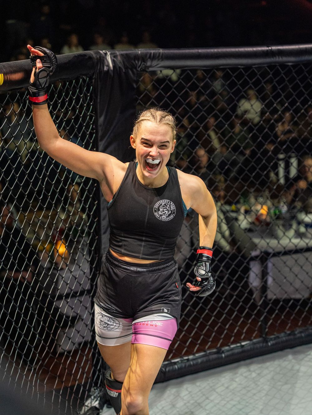 Female MMA fighter celebrates victory in cage, raising fist triumphantly, wearing black tank top and pink shorts.
