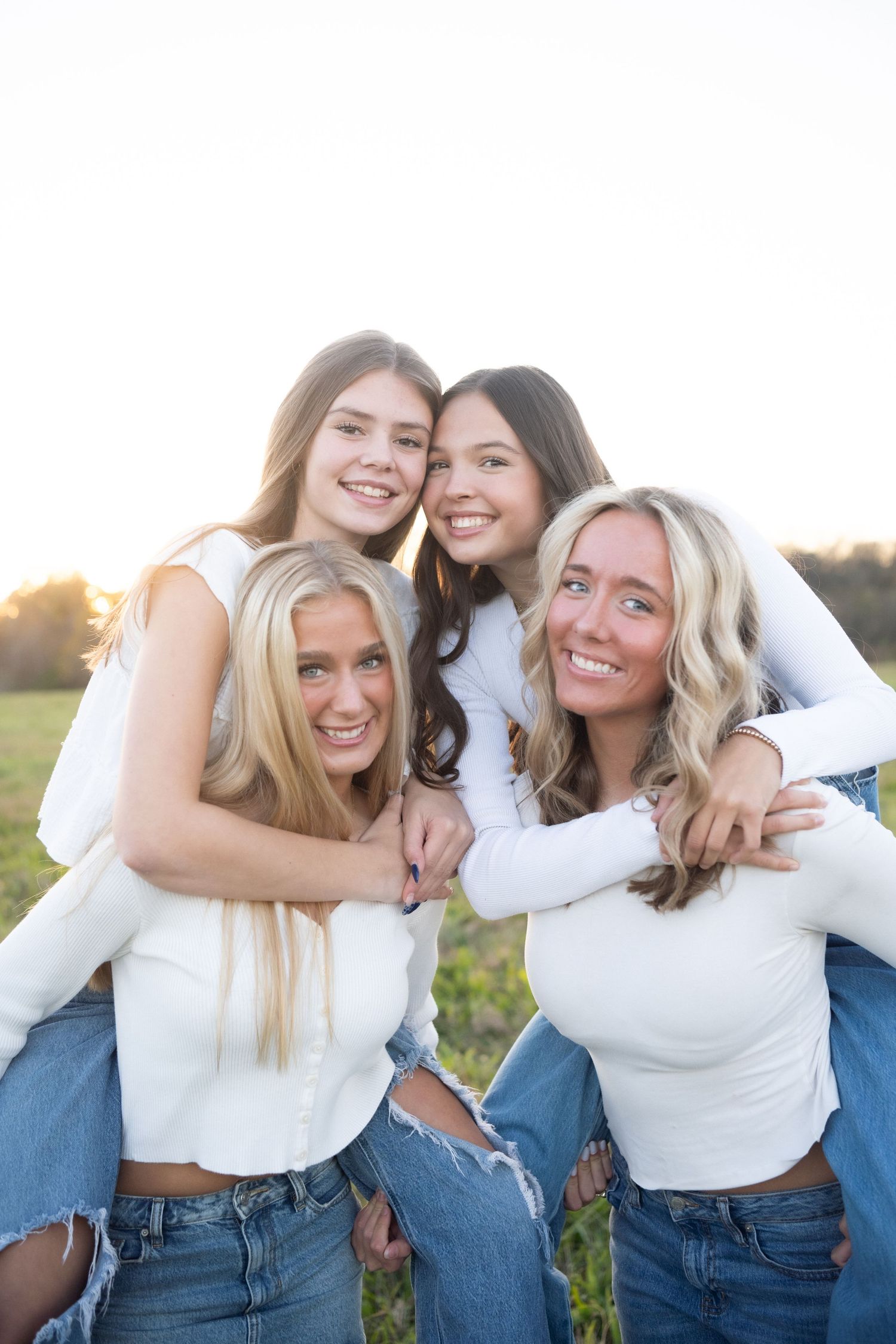 Group of friends wearing matching white tops and jeans pose together in a sunny field at sunset.