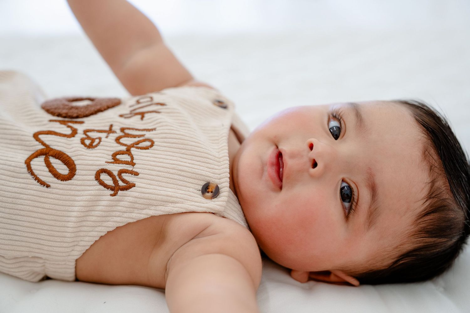 A baby wearing a beige and brown striped onesie lies on a white surface.