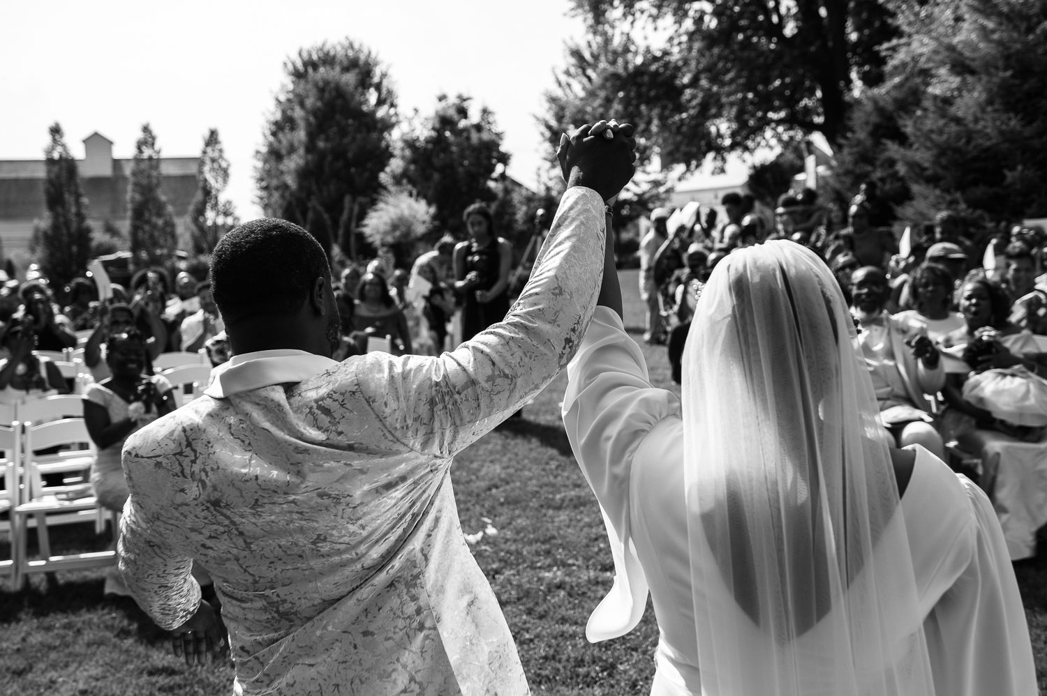 Black and white photo of a bride and groom raising hands in celebration at an outdoor wedding ceremony.
