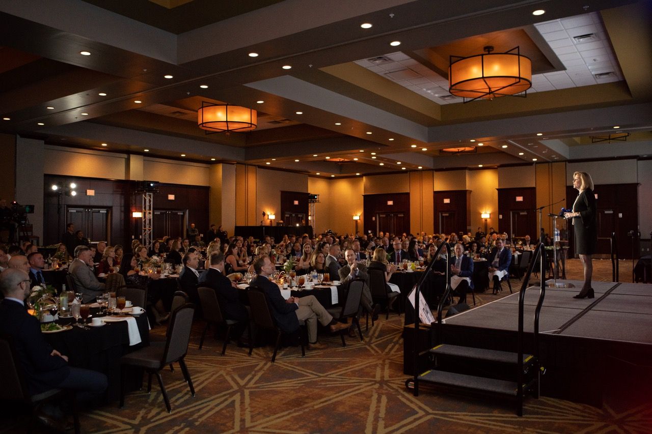 Large corporate banquet event in a hotel ballroom with a speaker on stage addressing hundreds of seated guests.