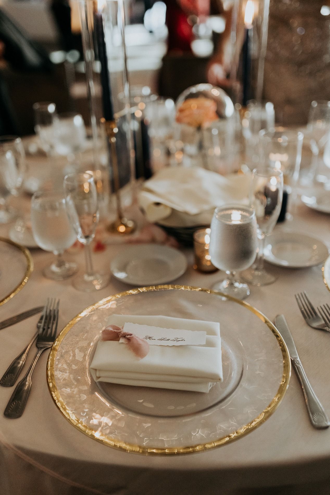Elegant wedding reception table setting with gold-rimmed charger plates, white napkins, crystal glassware, and tall black candles.