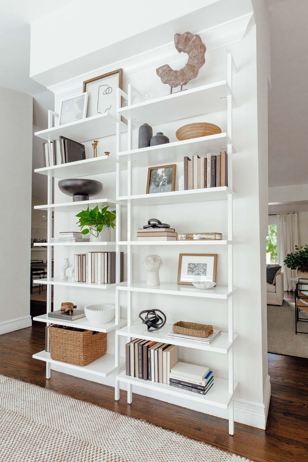 Tall white open bookshelf styled with neutral books, vases, plants, and woven baskets.