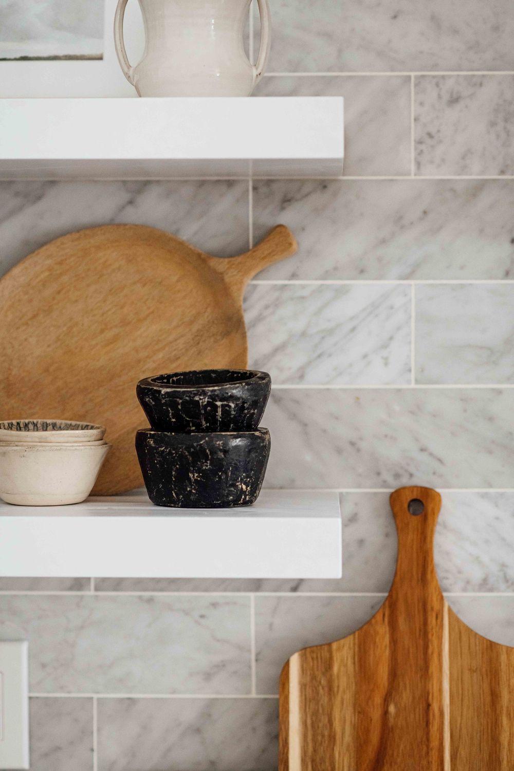 White floating kitchen shelves with wooden cutting boards and black bowls on marble tile backsplash.