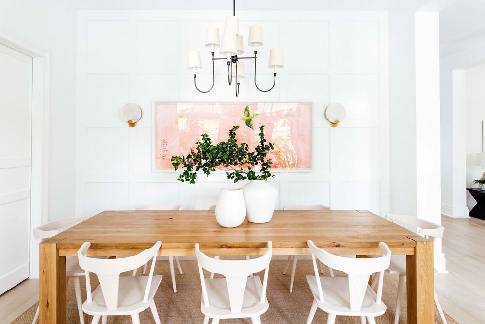 Bright dining room with wood table, white Windsor chairs, black chandelier, wall sconces, and pink floral artwork.