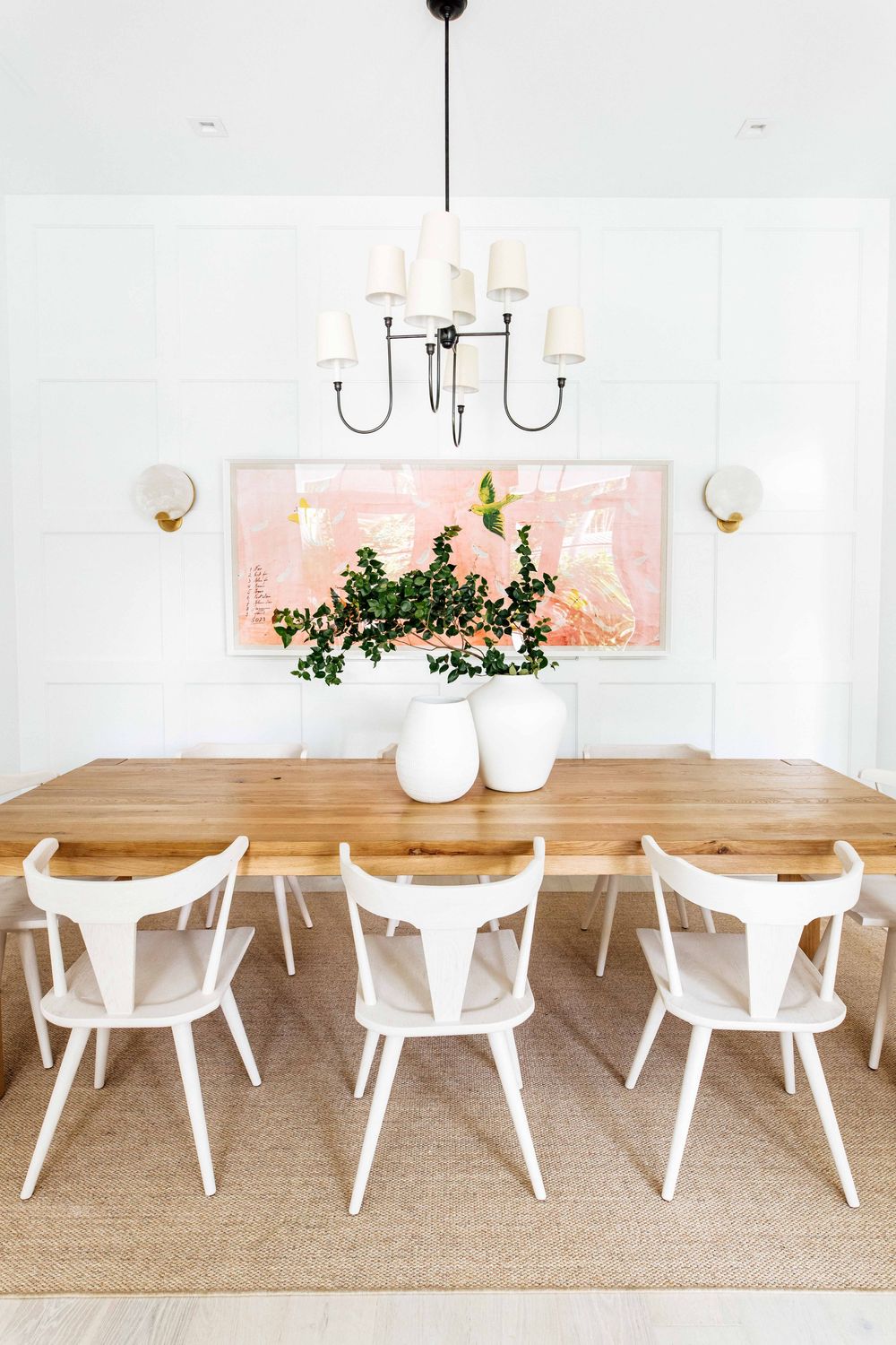 Elegant dining room with natural wood table, white chairs, black chandelier, jute rug, and large pink abstract artwork.