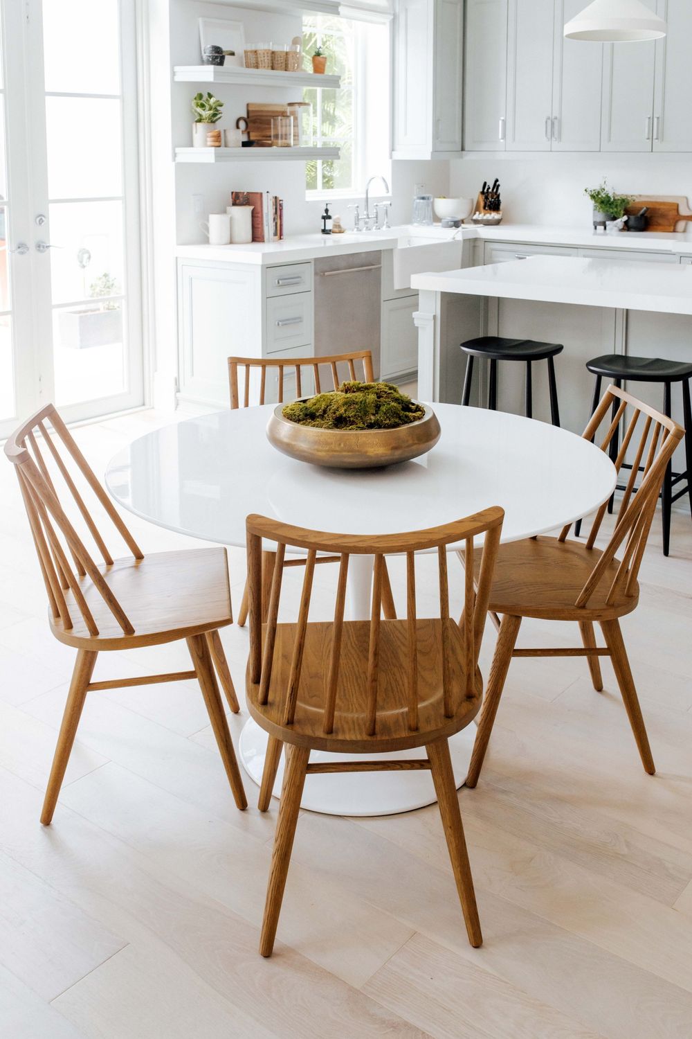 Modern white kitchen with round tulip dining table, natural wood Windsor chairs, and open shelving in background.