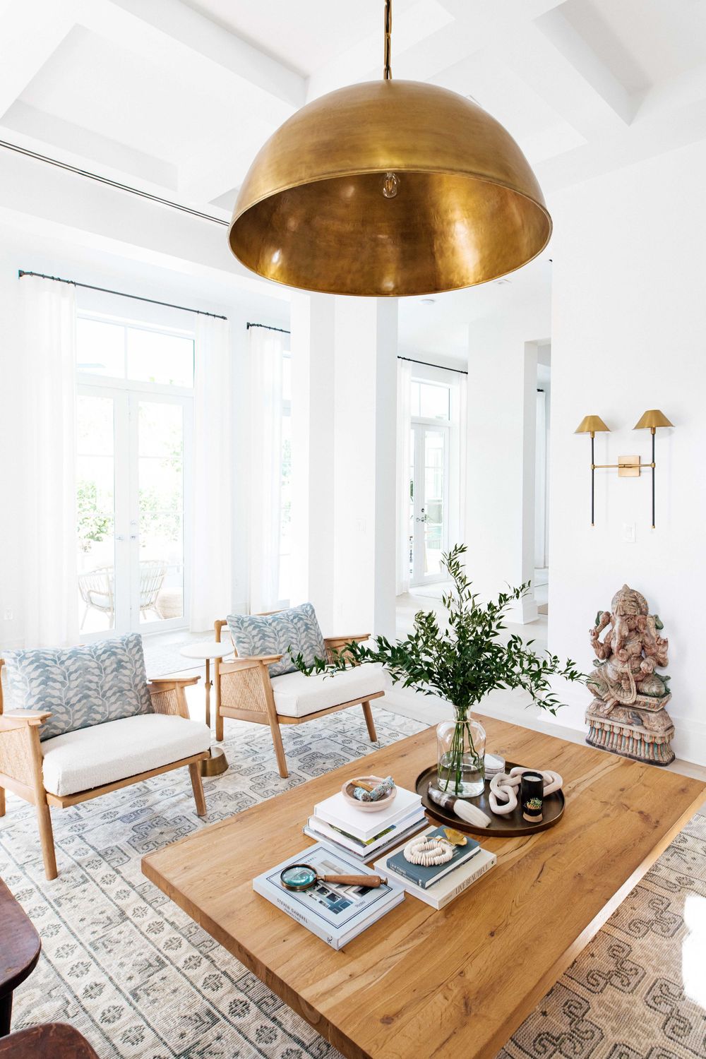 Airy living room featuring a brass dome pendant light, wooden chairs with blue pillows, and a rustic coffee table with books.