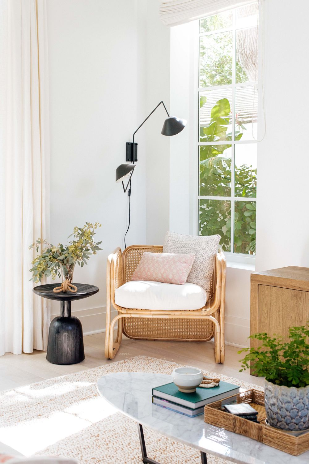 Bright corner nook with wicker rattan armchair, black swing arm wall lamp, small black side table, and tall windows.