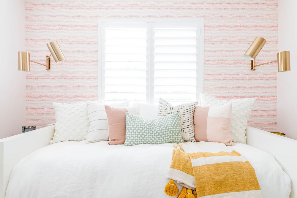 Cozy white daybed with pink and gray pillows against pink striped wallpaper with gold wall sconces.