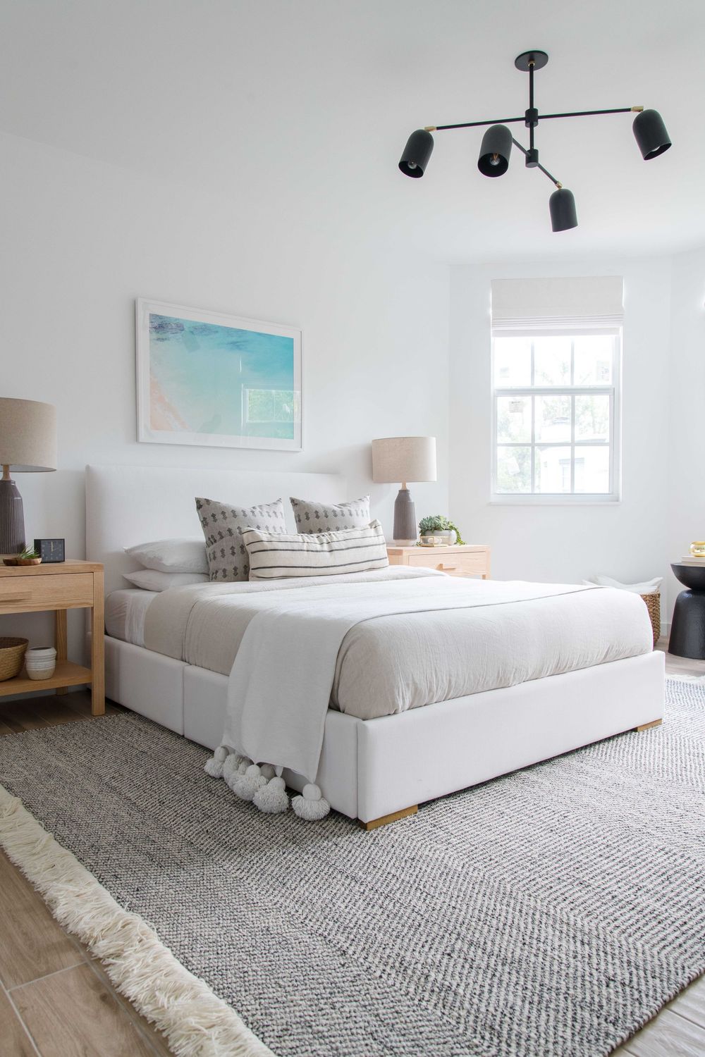 Serene white primary bedroom with upholstered bed, layered rugs, and aqua abstract wall art.