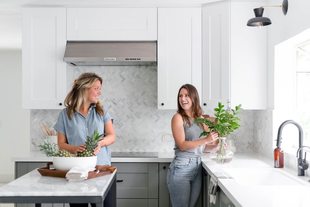 Two women arranging greenery in a modern white and gray kitchen with marble backsplash.
