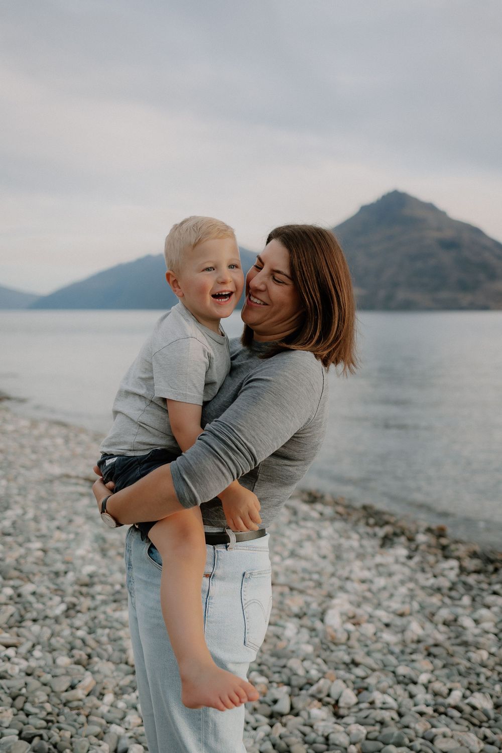 A mother holds her laughing toddler at Lake Wakatipu, Homestead Bay in Queenstown during a family photoshoot.
