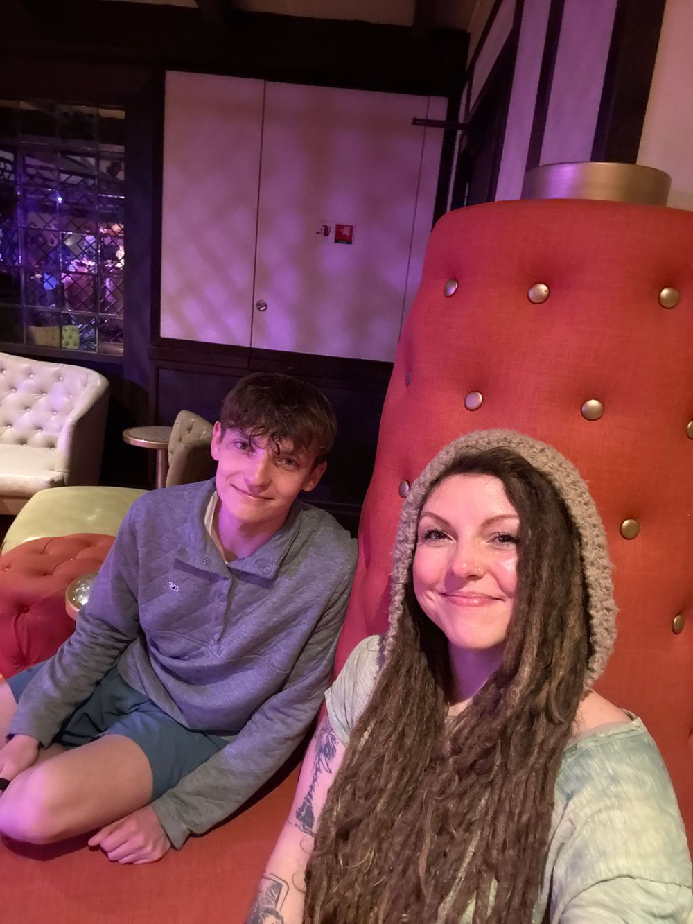 Two people sitting on colorful tufted chairs in a lounge, one wearing a knit hood with dreadlocks and a tattoo.