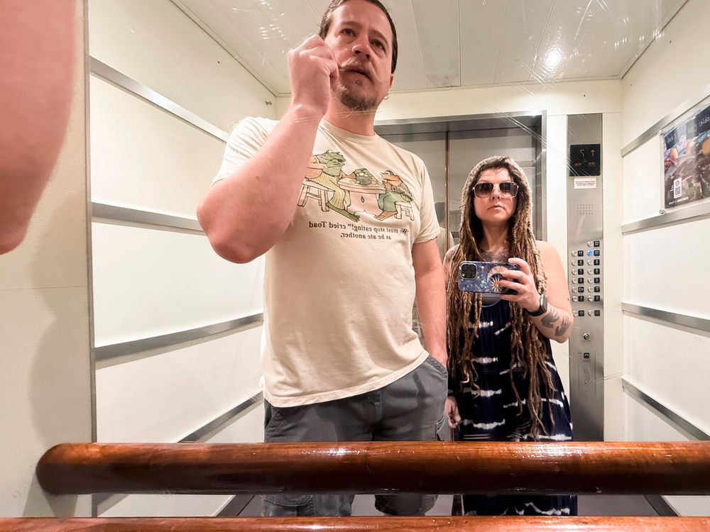 A man and woman taking a mirror selfie in an elevator, man raising hand, woman holding phone wearing sunglasses.