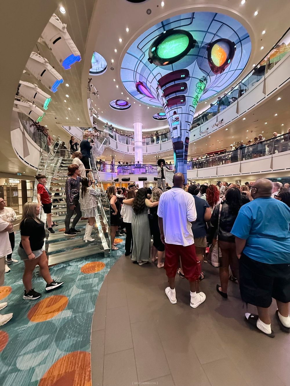 Crowded cruise ship atrium with colorful ceiling lights, multiple decks, and passengers gathered in a modern interior space.