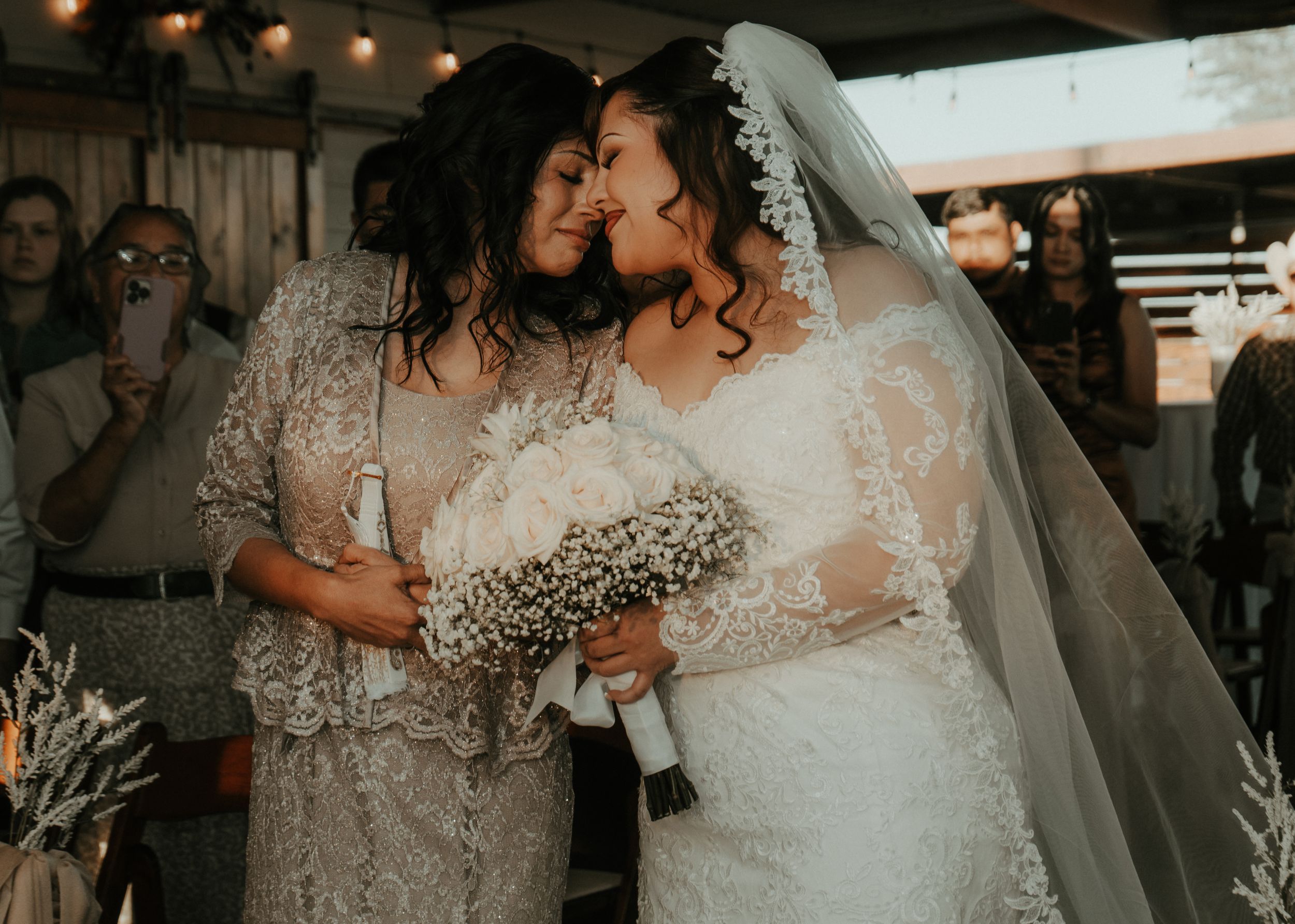 Two brides in white lace wedding gowns share an intimate moment, touching foreheads while holding bouquets at their wedding ceremony.