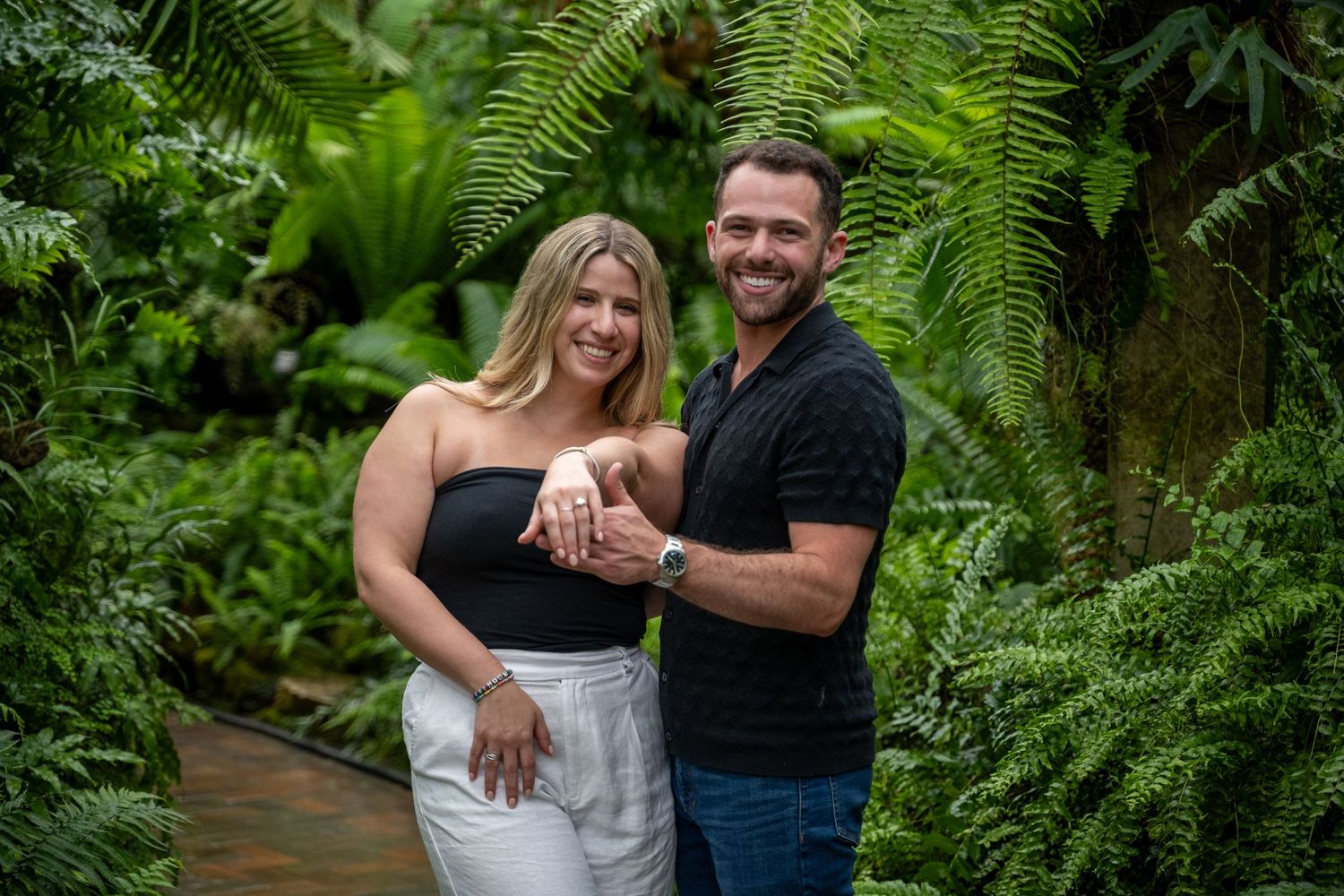 A couple shares a romantic moment surrounded by lush green ferns in a forest setting during an outdoor photo session.