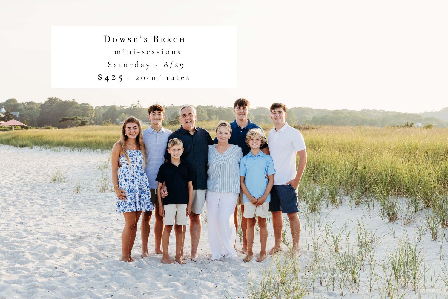 A group of people in coordinated white and blue outfits pose together on a beach with coastal grass at sunset.