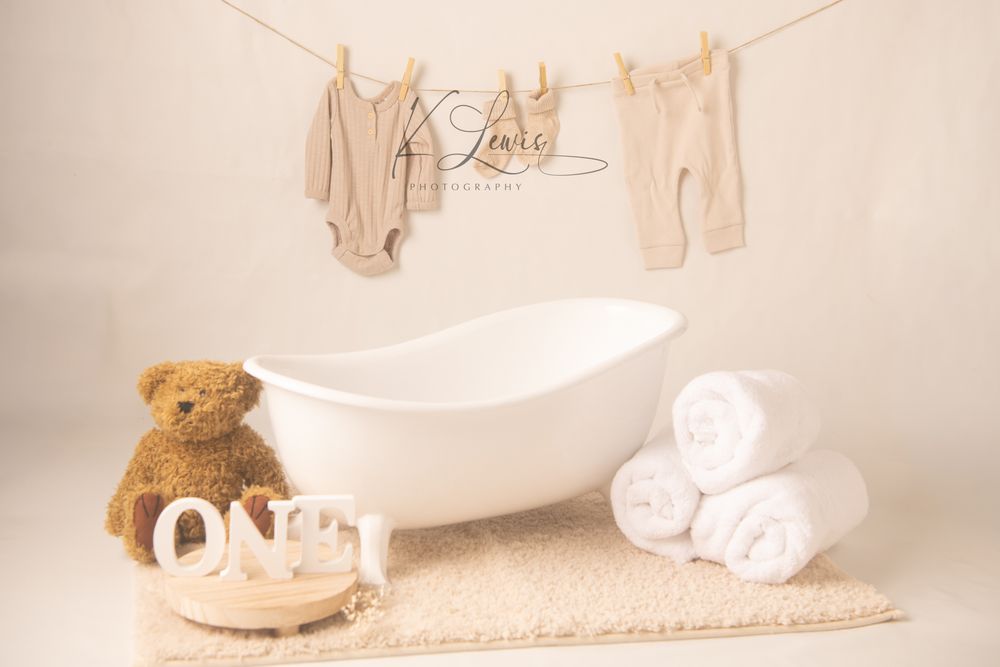Neutral toned newborn photo setup with white bathtub, teddy bear, rolled towels, and baby clothes on a clothesline.