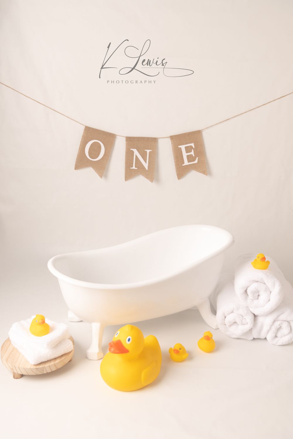White clawfoot tub with rubber ducks and rolled towels under a burlap ONE banner for a first birthday photo session.
