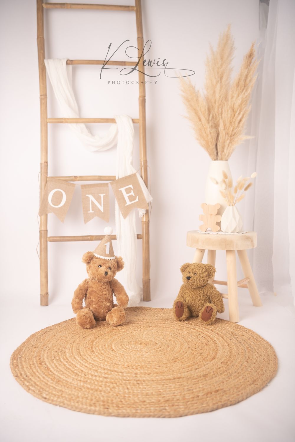 Two teddy bears sit on a round jute rug beside a wooden ladder with ONE banner and pampas grass decor.