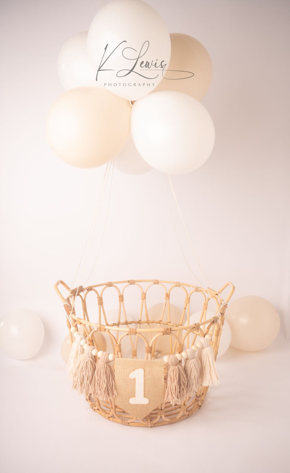Wicker basket with number 1 banner and cream tassels, suspended by neutral white balloons on a soft white background.