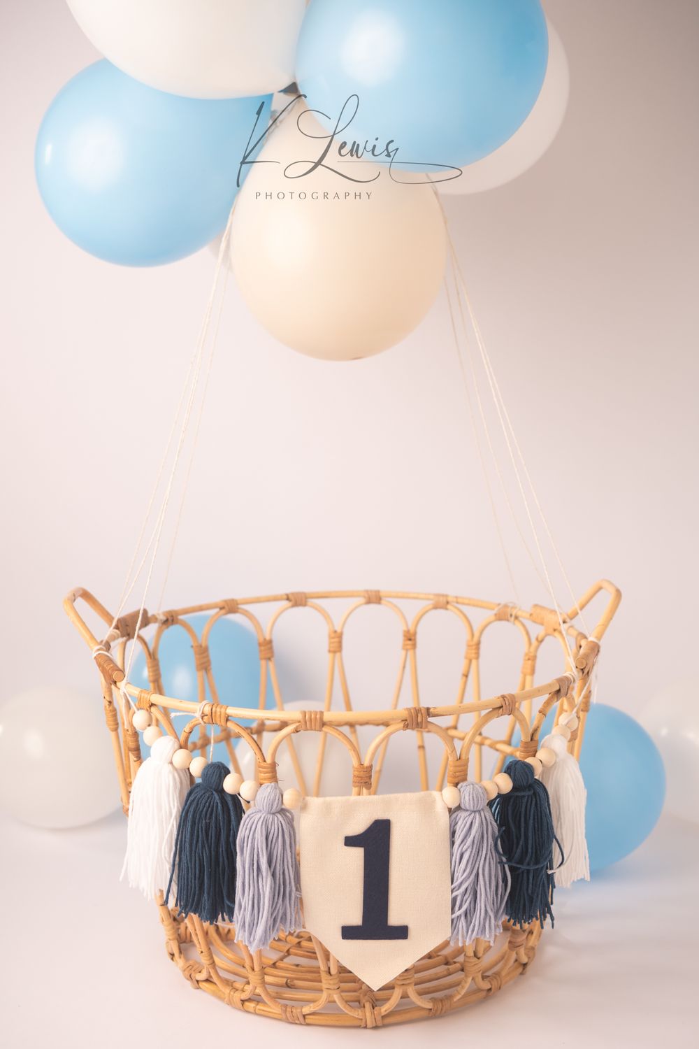Wicker basket with number 1 banner and blue tassels, suspended by blue and white balloons on white background.
