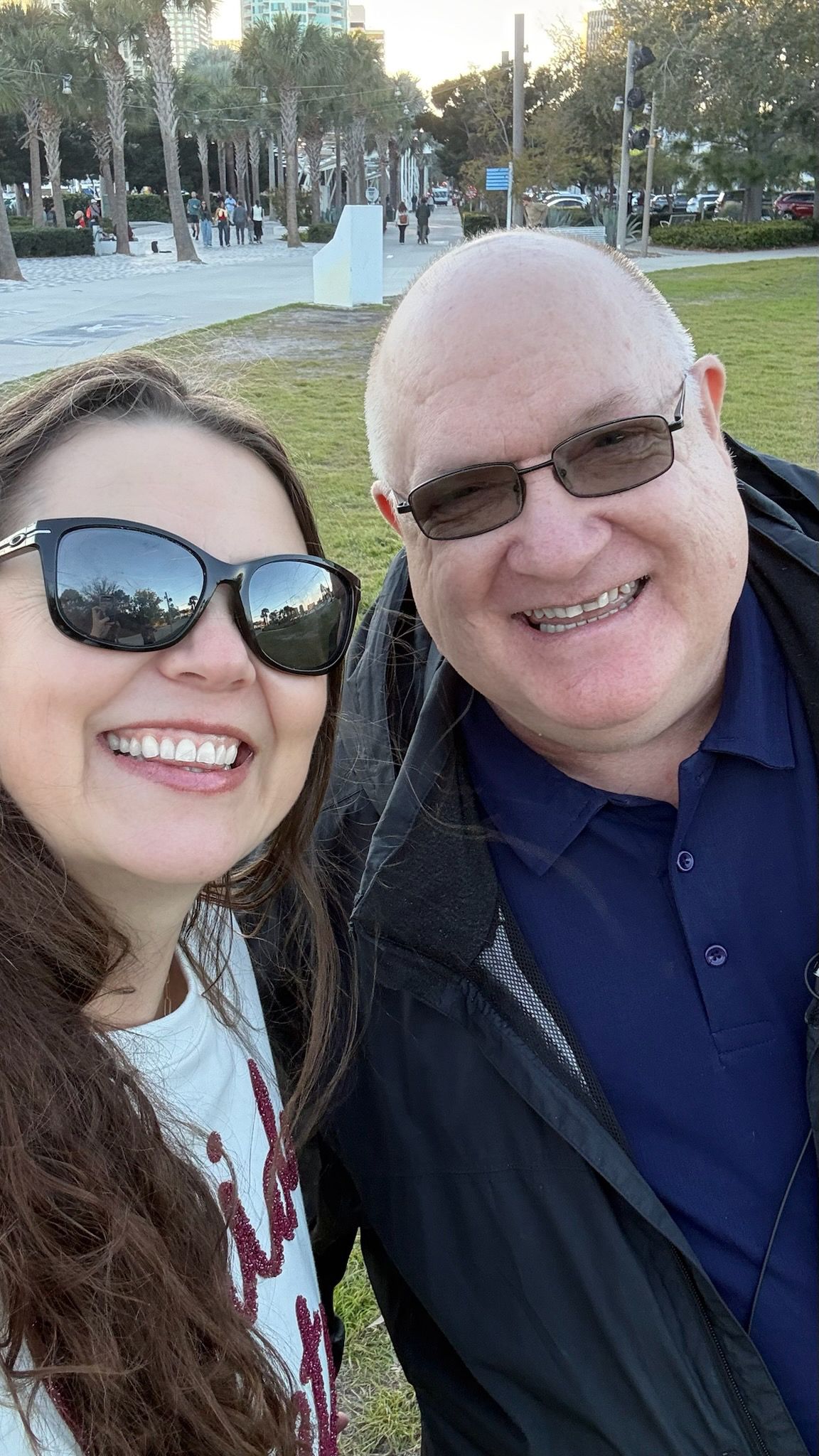 A smiling couple takes a selfie outdoors in a sunny park, both wearing sunglasses and casual clothing.