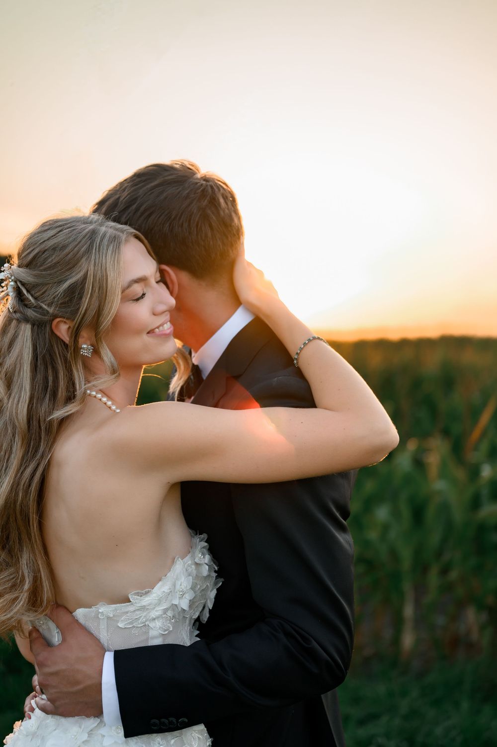 A bride and groom embrace at golden hour sunset, with the smiling bride in a white gown and the groom in a black suit.