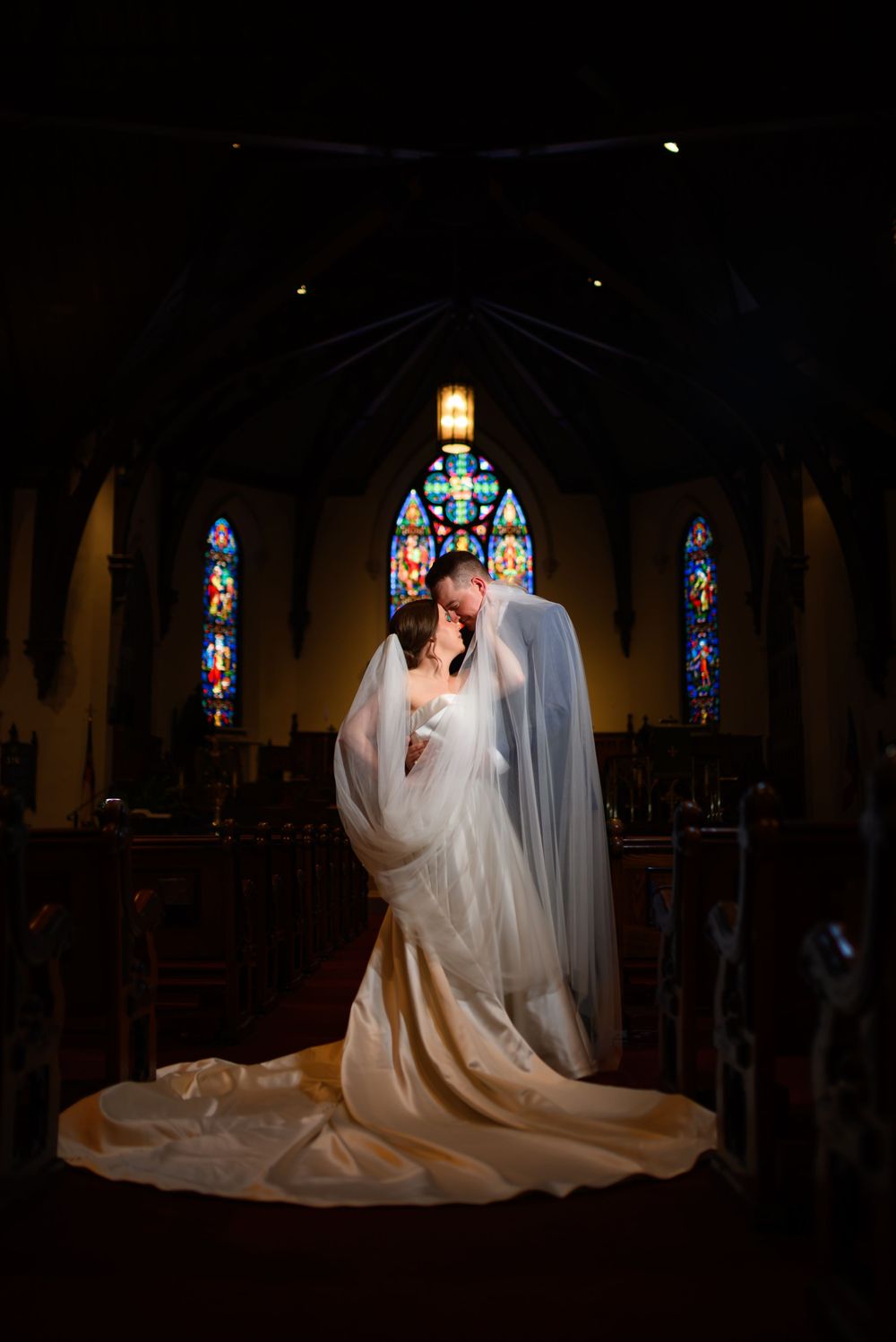 Bride and groom share a romantic kiss inside a church with stained glass windows and glowing altar lights.