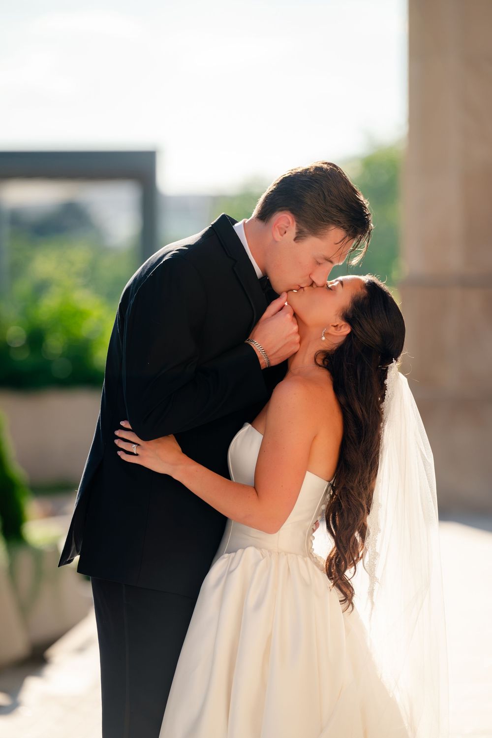 A bride and groom share a romantic kiss outdoors, with the bride in a strapless white gown and veil.