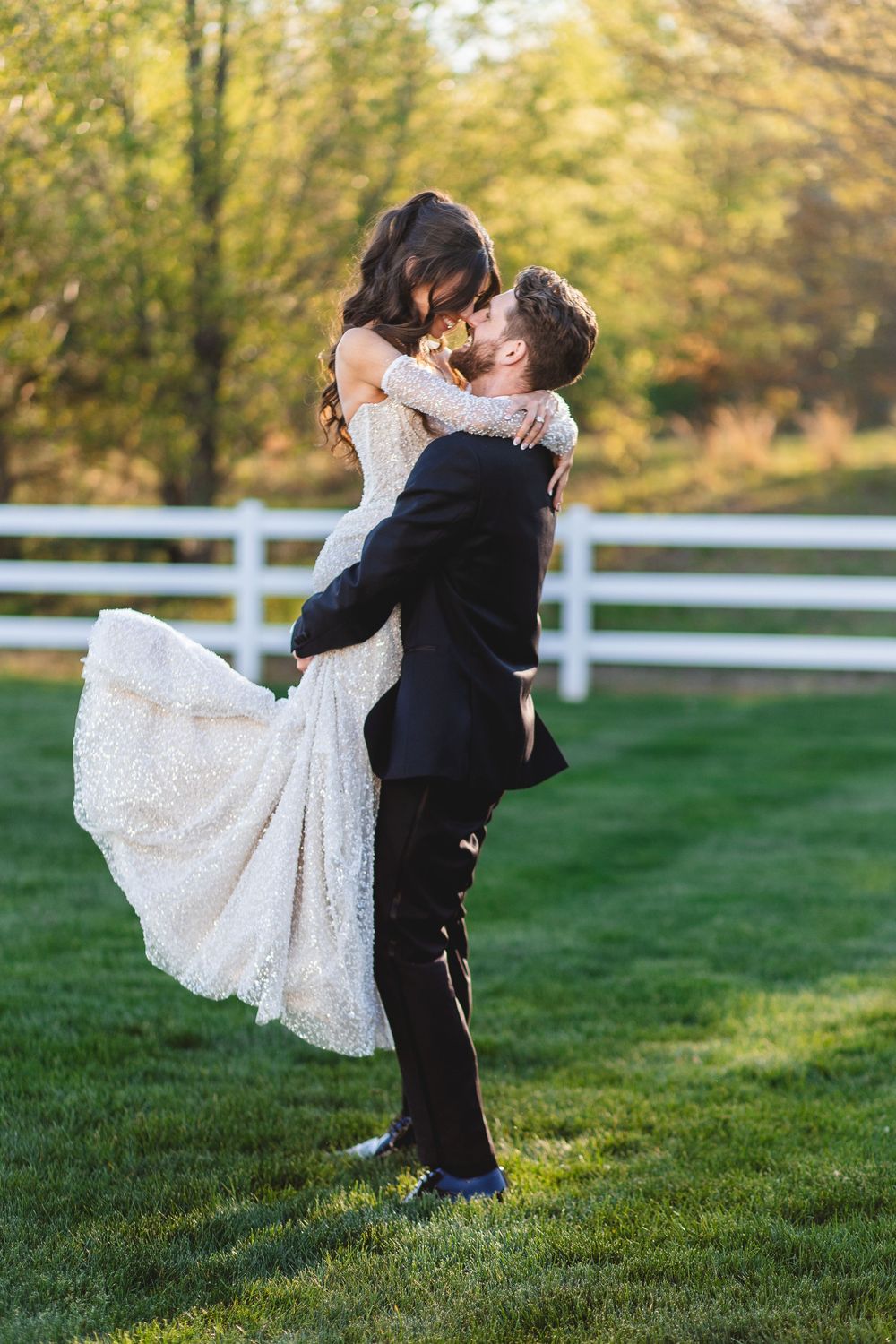 Groom lifts bride in lace wedding dress for a romantic kiss on green lawn with white fence backdrop.