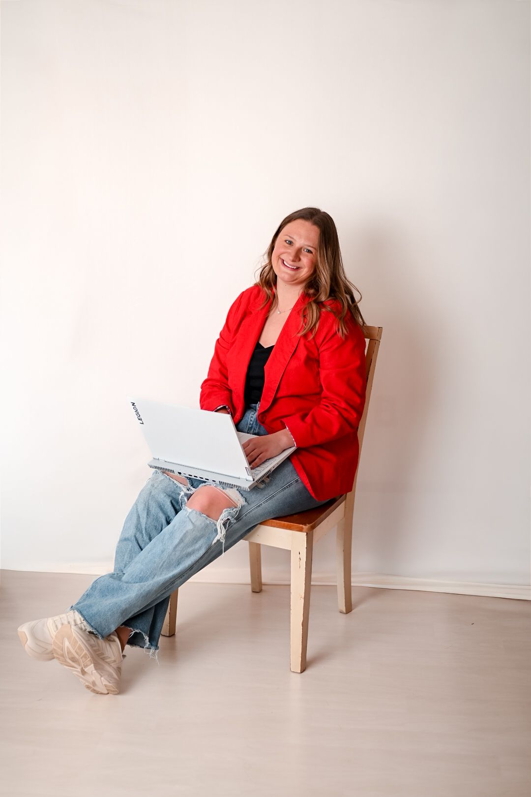 Smiling woman in red blazer and jeans sitting on wooden chair with laptop in bright room.