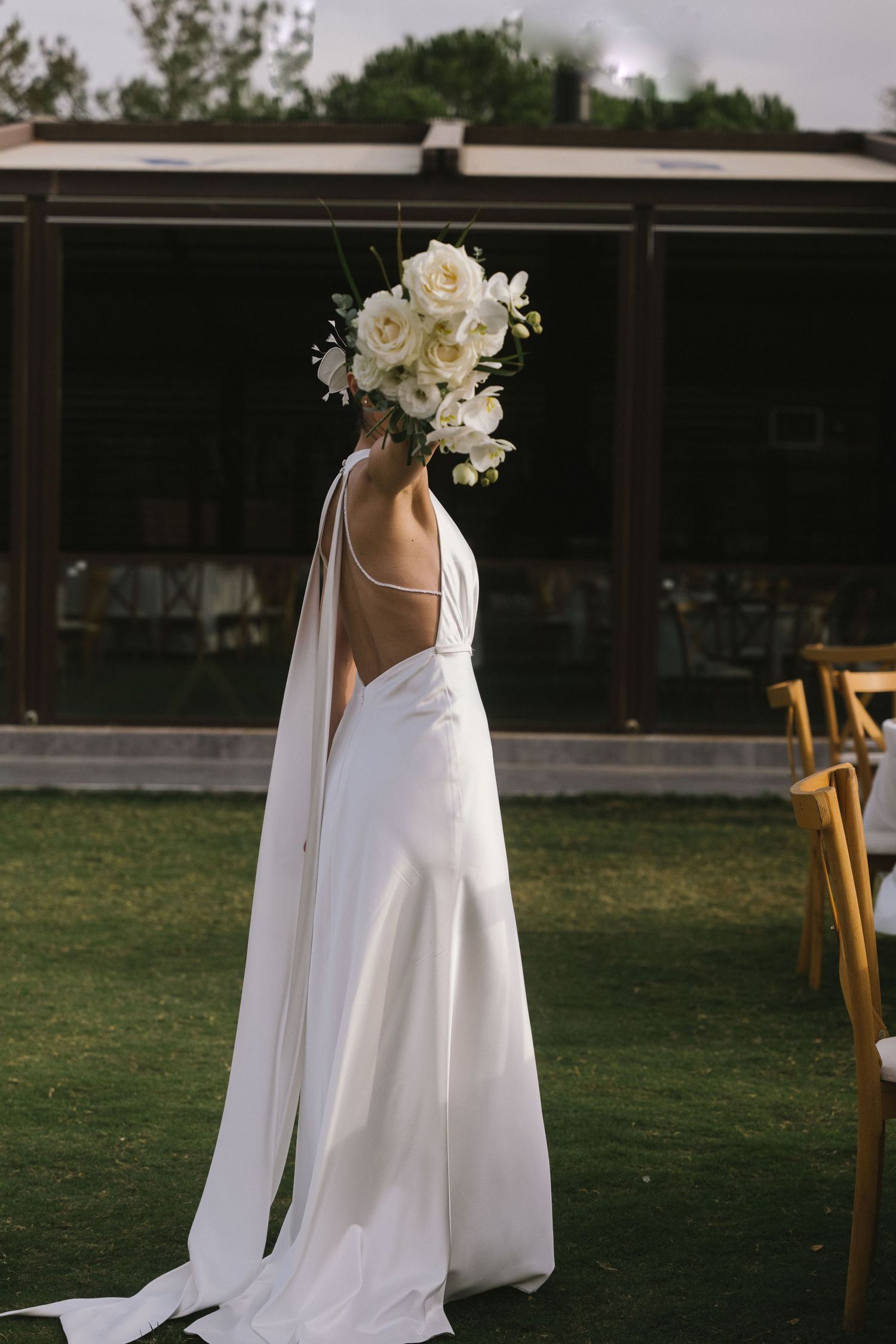 A bride in a flowing white wedding dress with cape details walks across a grassy lawn at an outdoor venue.