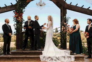 Wedding ceremony taking place under a rustic wooden pavilion decorated with floral arrangements at sunset.