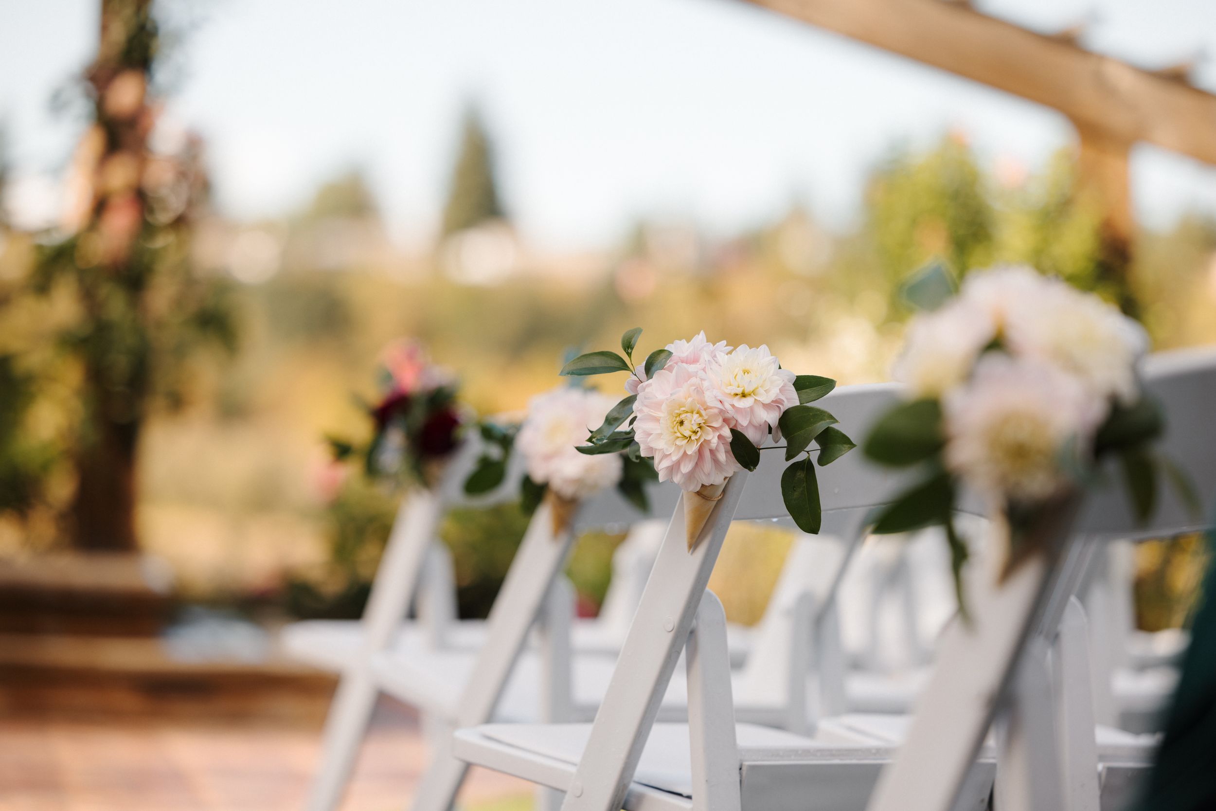 White folding chairs adorned with delicate pink floral arrangements line up for an outdoor wedding ceremony.