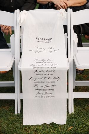 Reserved seating signs printed in elegant calligraphy on white paper hang from white chairs at an outdoor wedding ceremony.