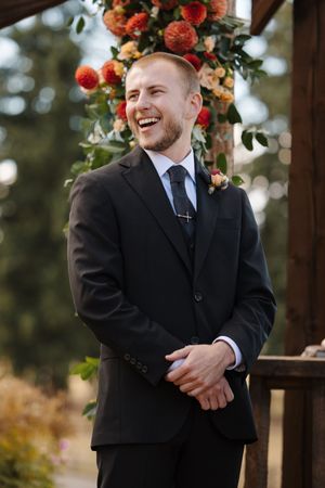 Groomsman in black suit smiling under wooden arch decorated with coral and peach colored floral arrangements.