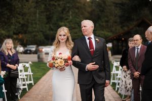 Wedding guests gather outdoors for a ceremony with white chairs arranged on a garden path.