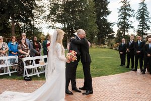 Outdoor wedding ceremony with guests seated in white chairs surrounded by evergreen trees.