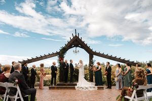 A panoramic view of an outdoor wedding ceremony under a wooden arch with mountain vistas and cloudy skies in the background.