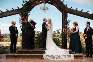 A wedding ceremony takes place under a rustic wooden pergola with flowers and greenery during a beautiful outdoor celebration.