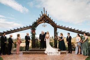 Wedding ceremony takes place under a wooden pergola structure overlooking a scenic coastal landscape at sunset.