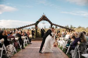 A couple shares their first kiss under a rustic wooden arch at an outdoor wedding ceremony surrounded by guests.