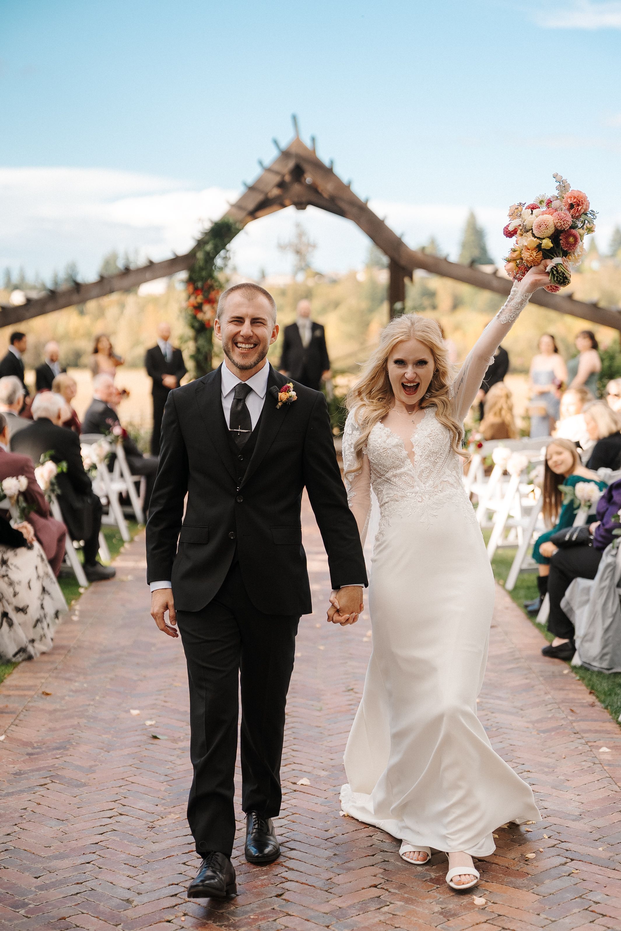 A joyful couple walks down a brick pathway during their outdoor wedding ceremony while guests look on.